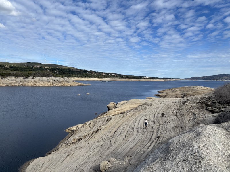 A small figure stands on the banded shore, with blue water to the left and wind turbines on the distant ridge.
